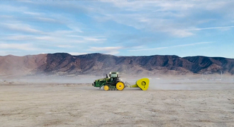 Green tractor with a large yellow implement working on a dusty, barren desert plain with mountains in the distance.