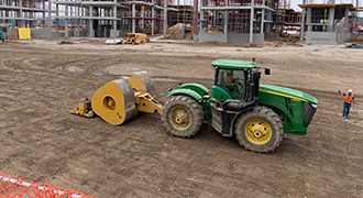 Green John Deere tractor pulling a yellow soil compactor on a dirt construction site, with buildings and scaffolding in the background.