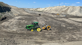 Green John Deere soil compactor on a dirt construction site with a yellow roller in the background, preparing the ground.