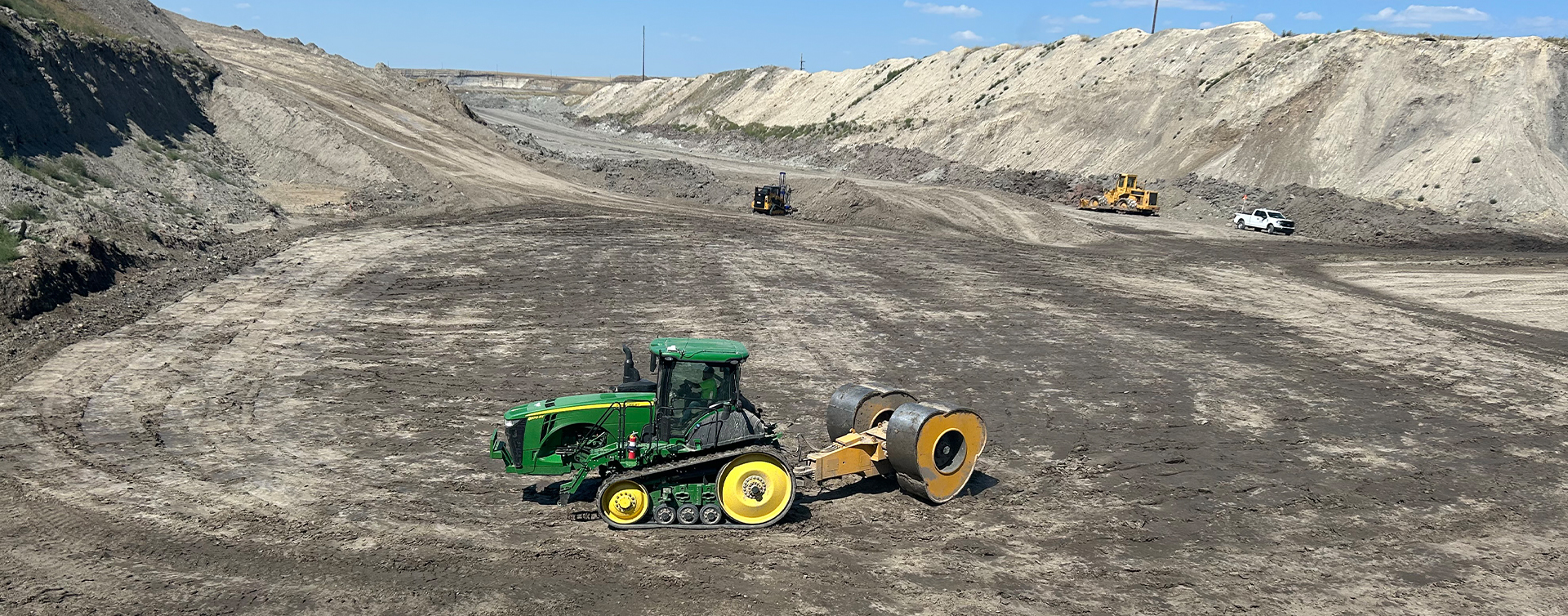 Green tracked bulldozer with a road roller attached, moving across a muddy construction site with dirt embankments in the background.